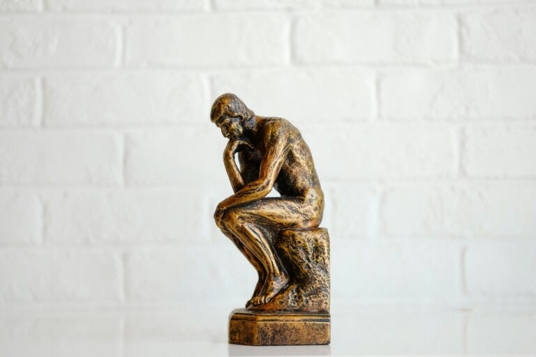 Bronzefarbene Skulptur von Auguste Rodins "Der Denker" auf einem Holzsockel vor einer weißen Ziegelmauer.
