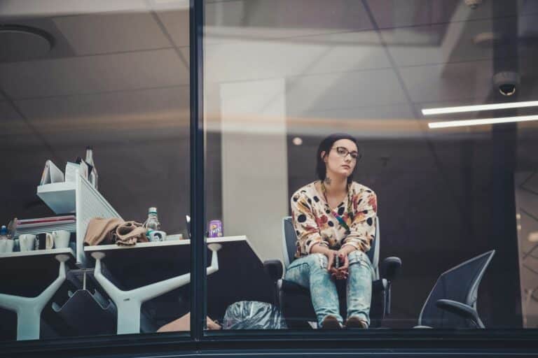 Frau sitzt nachdenklich im Büro, umgeben von Arbeitsutensilien, während sie aus dem Fenster blickt